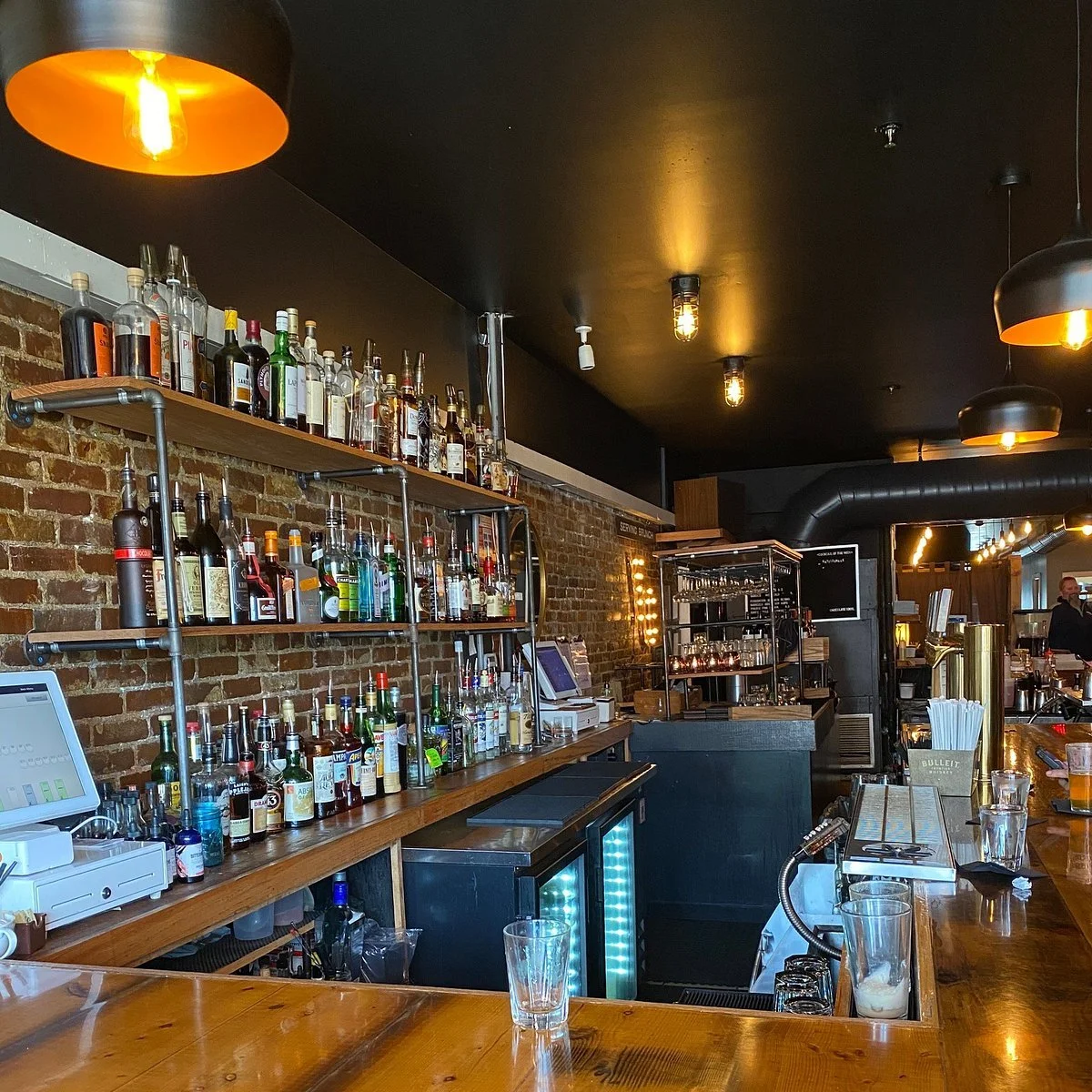 Interior of a bar with a brick wall, shelves filled with liquor bottles, and a wooden bar counter with glasses and bar tools.
