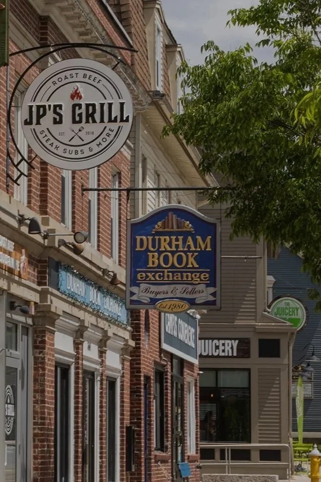 Street view of shops including JP's Grill restaurant, Durham Book Exchange, and a juicery, with brick and wooden buildings, signs, and green trees.