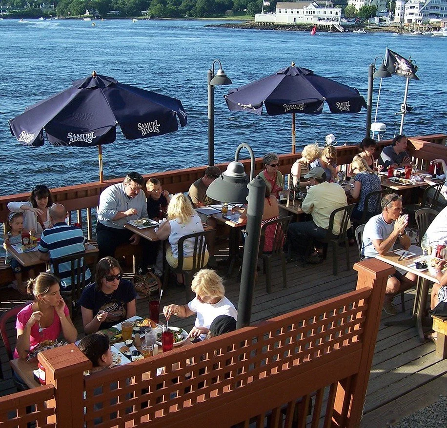 People dining outdoors on a patio by a river, with umbrellas, tables, and chairs.