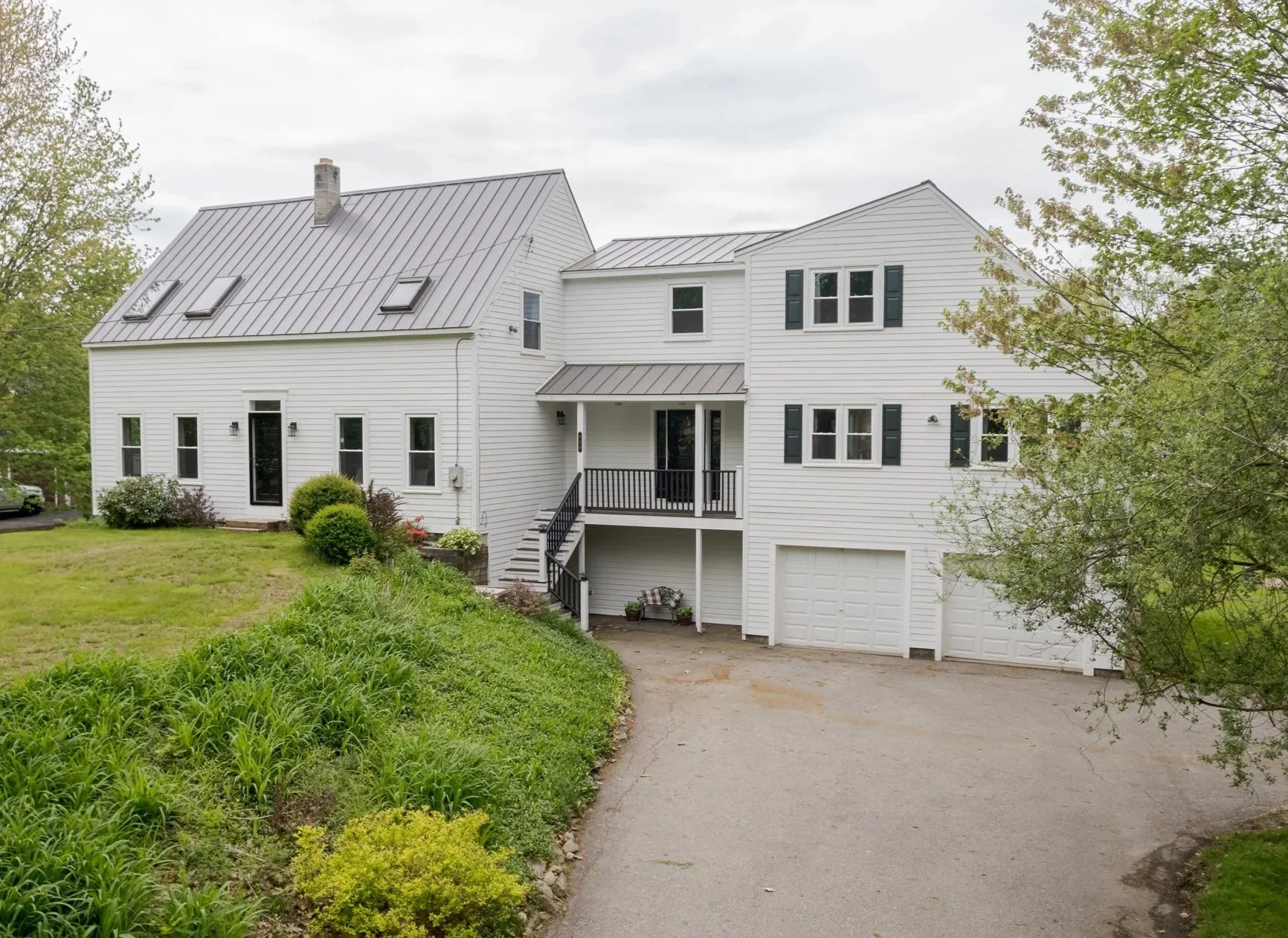 A modern two-story white house with black shutters, a metal roof, and a small front porch with stairs. The house has several windows, a garage door underneath the porch, and is surrounded by greenery and trees.