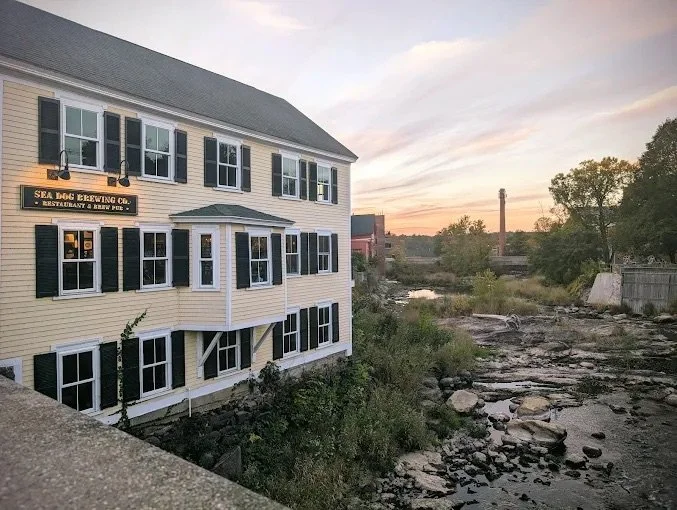 A white multi-story building with black shutters houses the Sea Bream Brewing Company restaurant and brew pub, situated next to a river or stream at sunset with trees and industrial buildings in the background.