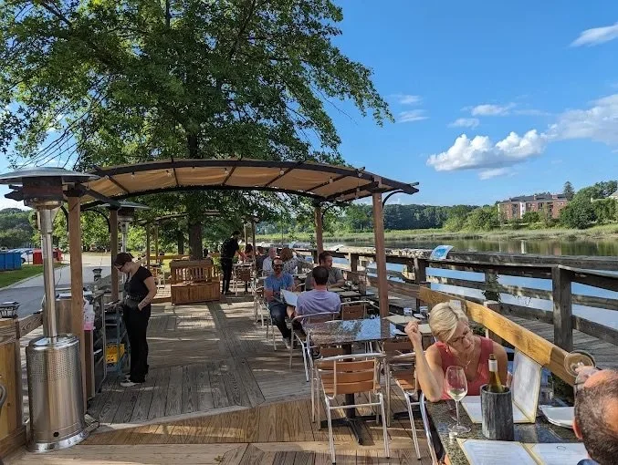 People dining and socializing on a wooden outdoor deck by a river, with trees and a blue sky in the background, shaded by an awning.