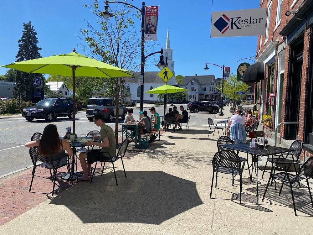 Outdoor café on a sunny day with people sitting at tables with green umbrellas on the sidewalk in a small town. A church with a steeple is visible in the background.