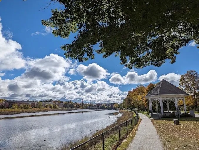 Scenic park with a river, a paved walkway, a white gazebo, trees with fall foliage, and a partly cloudy sky.