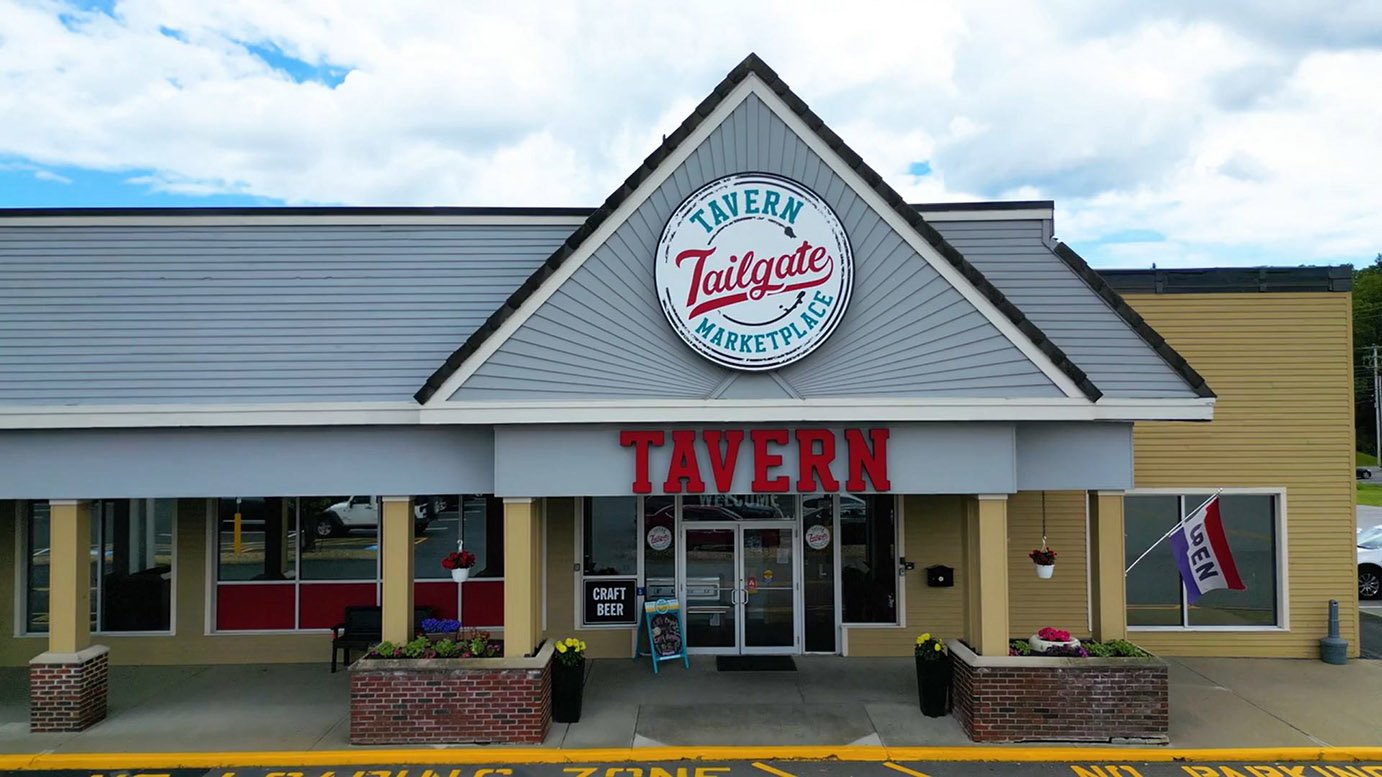 Exterior of a building with a sign reading 'Tavern Tailgate Marketplace,' with red 'Tavern' lettering above the entrance, a flag that says 'Menu,' and potted flowers on the sidewalk.