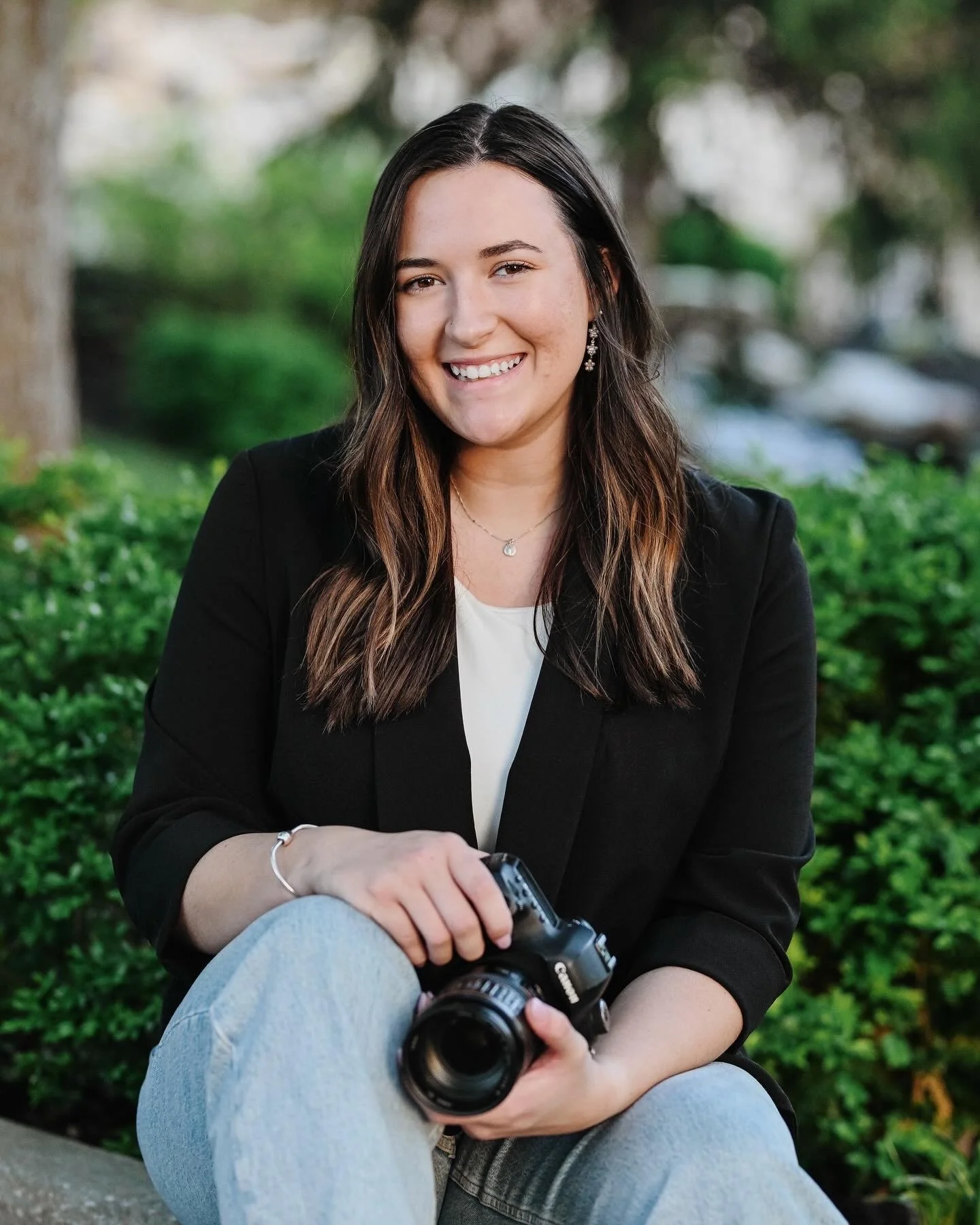 A young woman with long dark hair, smiling, sitting outdoors in a park, holding a camera, wearing a black blazer over a white shirt, with greenery in the background.