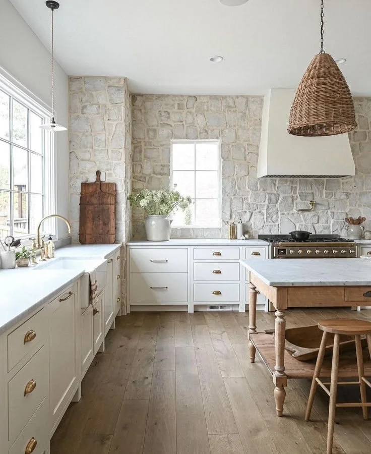 Modern kitchen with white cabinets, a stone wall, wooden kitchen table, wicker pendant light, and large window.