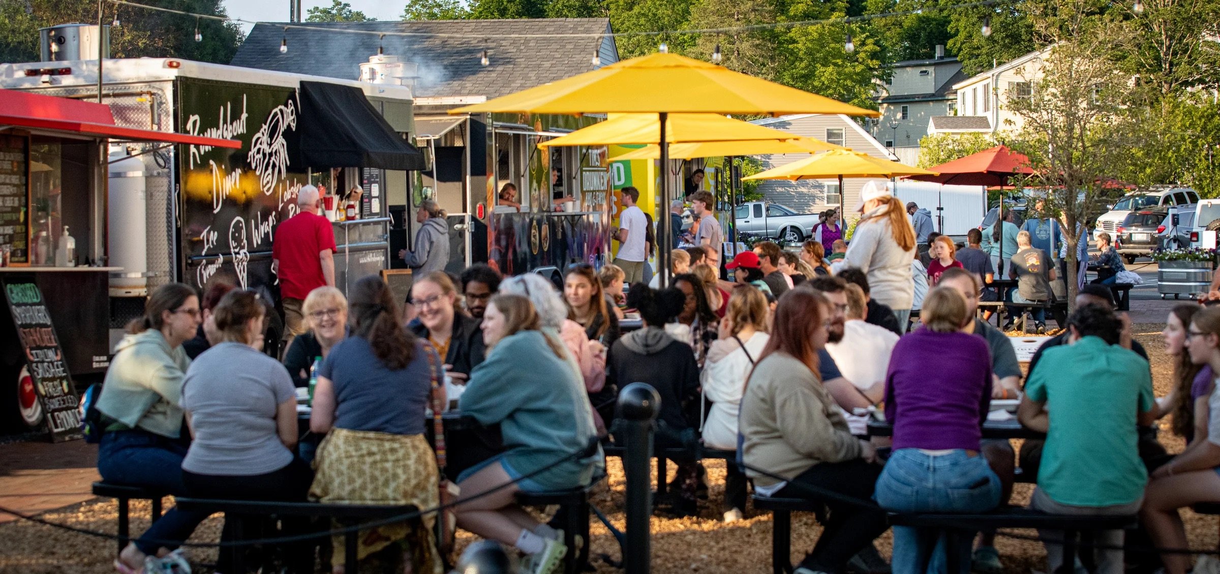 An outdoor food festival with many people sitting at tables and food trucks serving customers. Yellow umbrellas provide shade amongst trees and buildings.