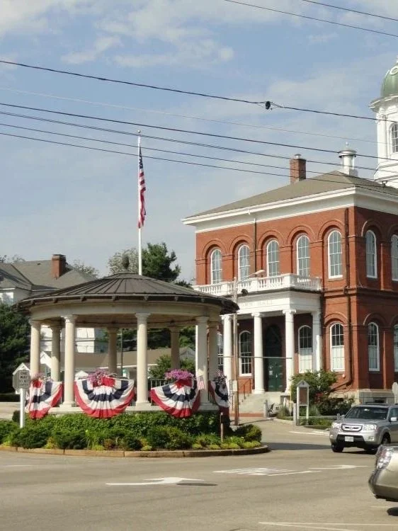 Red brick historic building with white trim and a dome, small white pavilion draped with American flag bunting, American flag flying on a pole, cars parked along the street, and a blue sky with few clouds.