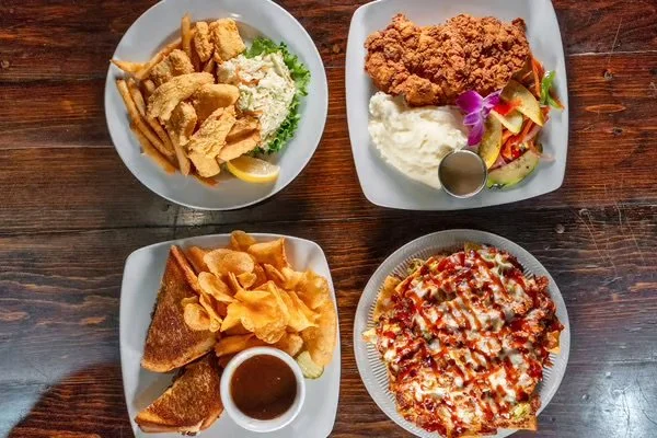 Four plates of food on a wooden table. Top left: fried fish fillets with French fries, coleslaw, and a lemon wedge. Top right: fried chicken breast with mashed potatoes, gravy, and a small serving of vegetables with an edible flower. Bottom left: grilled cheese sandwich with potato chips and a small bowl of dipping sauce. Bottom right: a pizza with tomato sauce, cheese, and toppings.