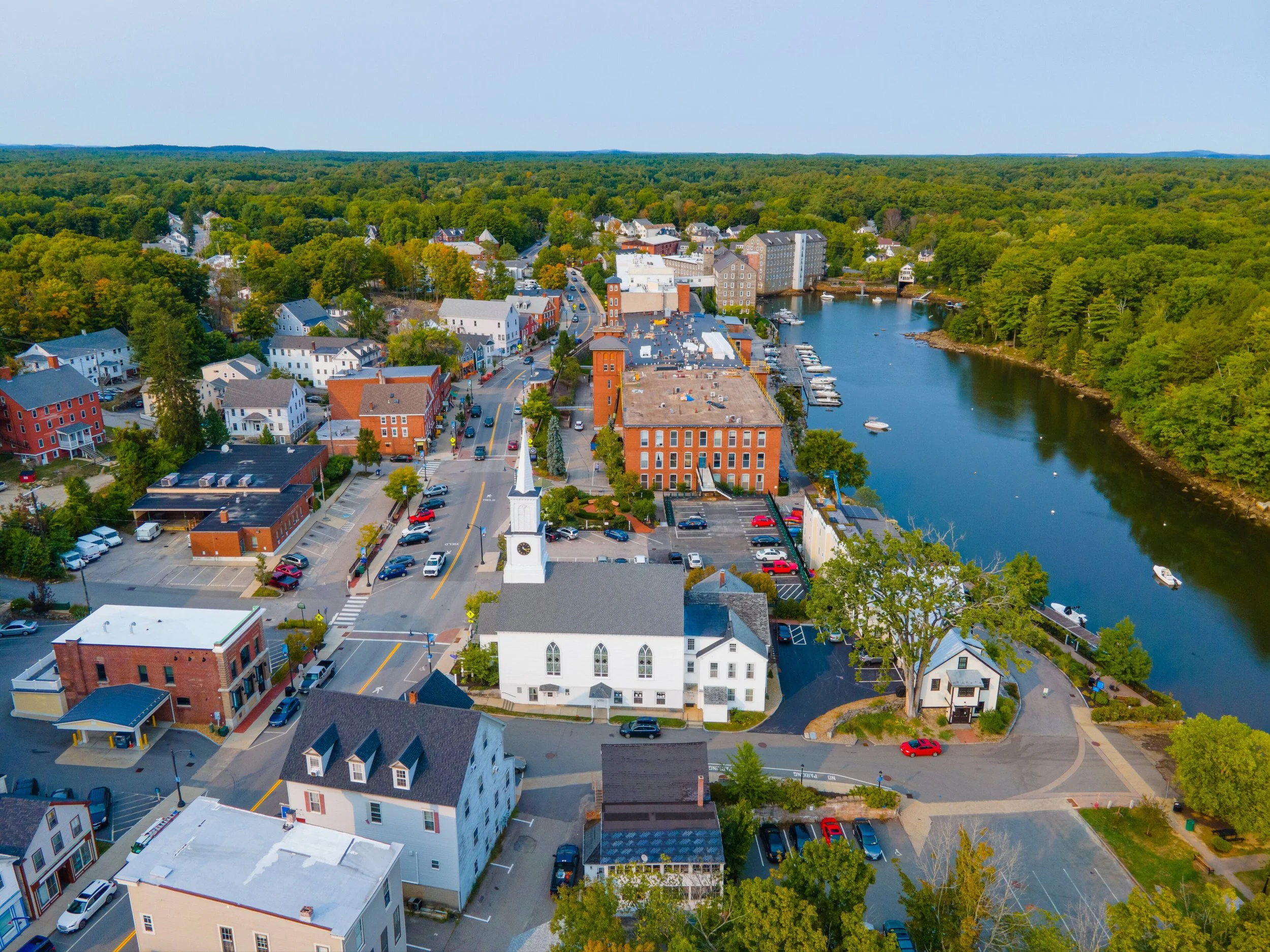Aerial view of a small town with colorful buildings, a river with boats, a clock tower, and a forested background.