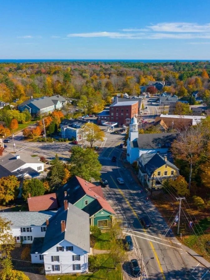 An aerial view of a small town with colorful houses, a church with a clock tower, and trees with fall foliage, stretching towards the horizon under a clear blue sky.