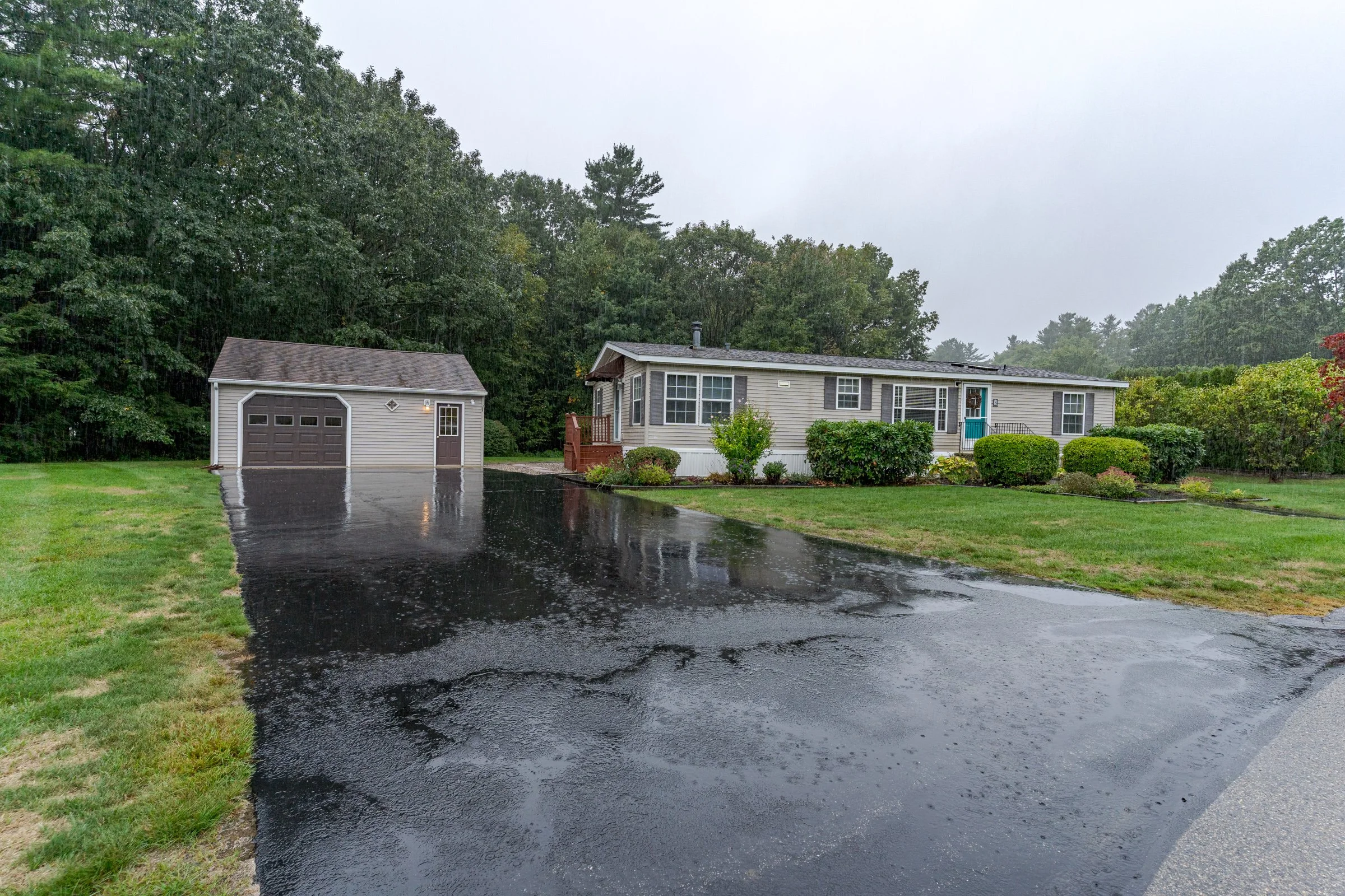 A house with a detached garage surrounded by trees, with a wet driveway and grass, during rain and overcast skies.