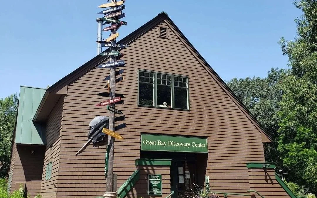 A wooden building with a green sign reading 'Great Bay Discovery Center', a window, and a flagpole with multiple directional signs.