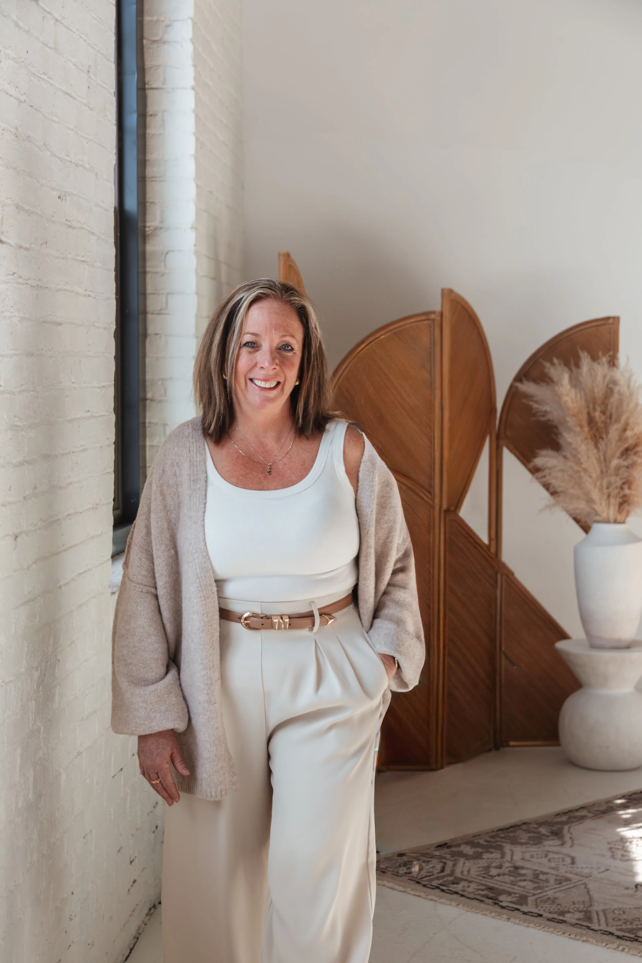 A woman with shoulder-length brown hair, wearing a white sleeveless top, beige cardigan, and high-waisted cream pants with a beige belt, standing indoors next to a white brick wall and wooden room divider with decorative pampas grass in a white vase.