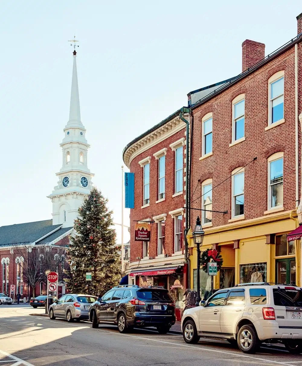 Street view of a small town with parked cars, brick buildings, a Christmas tree, a church steeple, and a clear sky.
