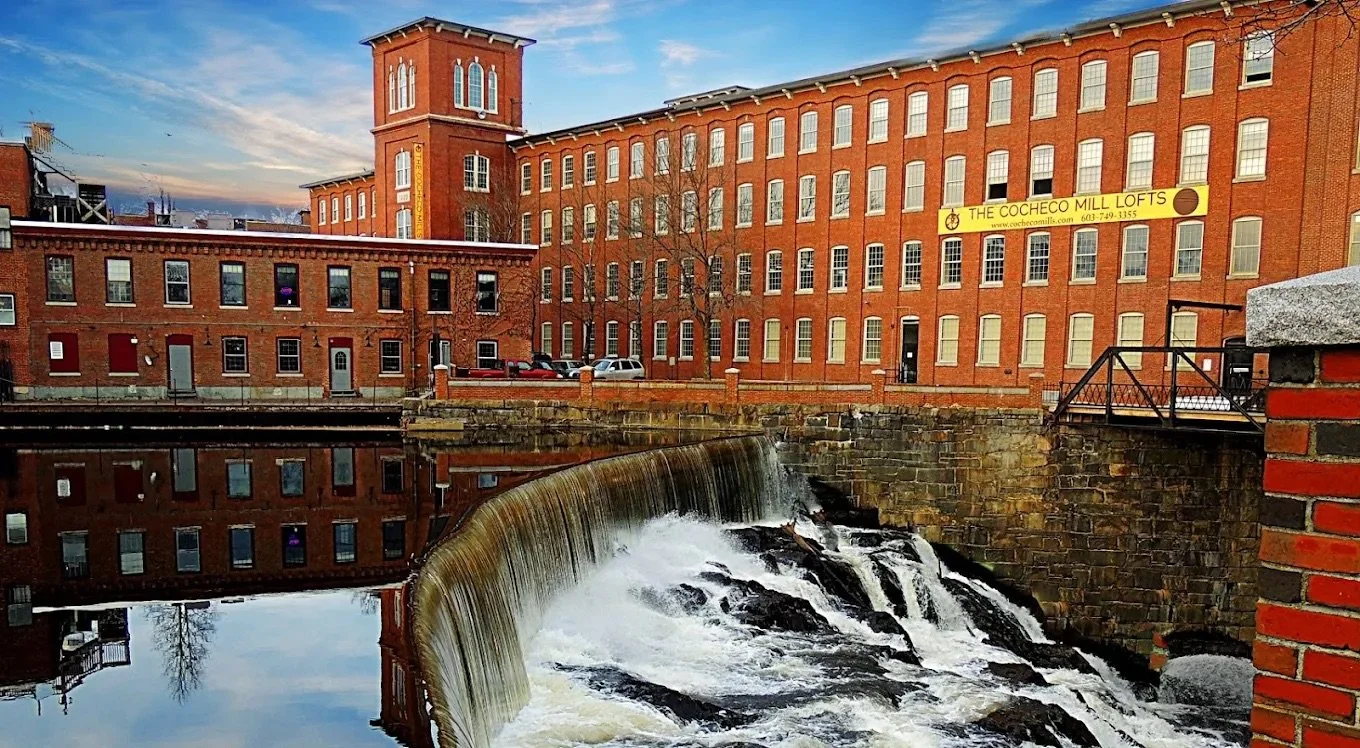 Historic red brick mill building next to a waterway with a small waterfall, reflecting in the water, under a partly cloudy sky.