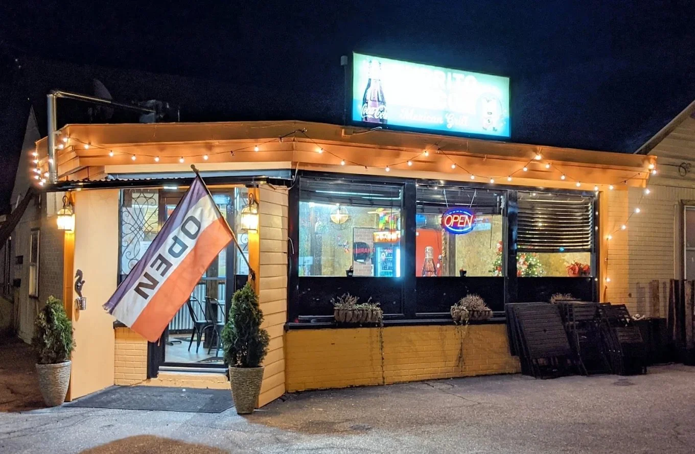 A small restaurant or cafe at night with a yellow exterior, string lights, and an orange and white flag that reads 'OPEN'. There are potted plants, a bench, and a large window with a neon 