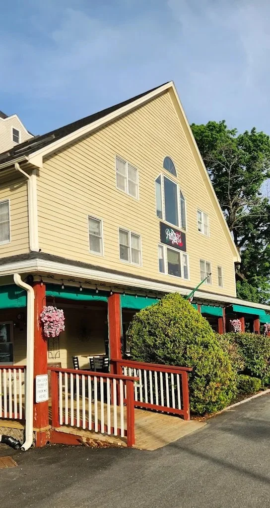A three-story building with yellow siding and multiple windows, including a rounded window near the peak of the roof. There is a sign that reads 'Wally's' on the upper part of the building. The lower level has an outdoor patio area with a green awning, red support beams, hanging flower baskets, and some chairs and tables, surrounded by bushes and a wooden railing.