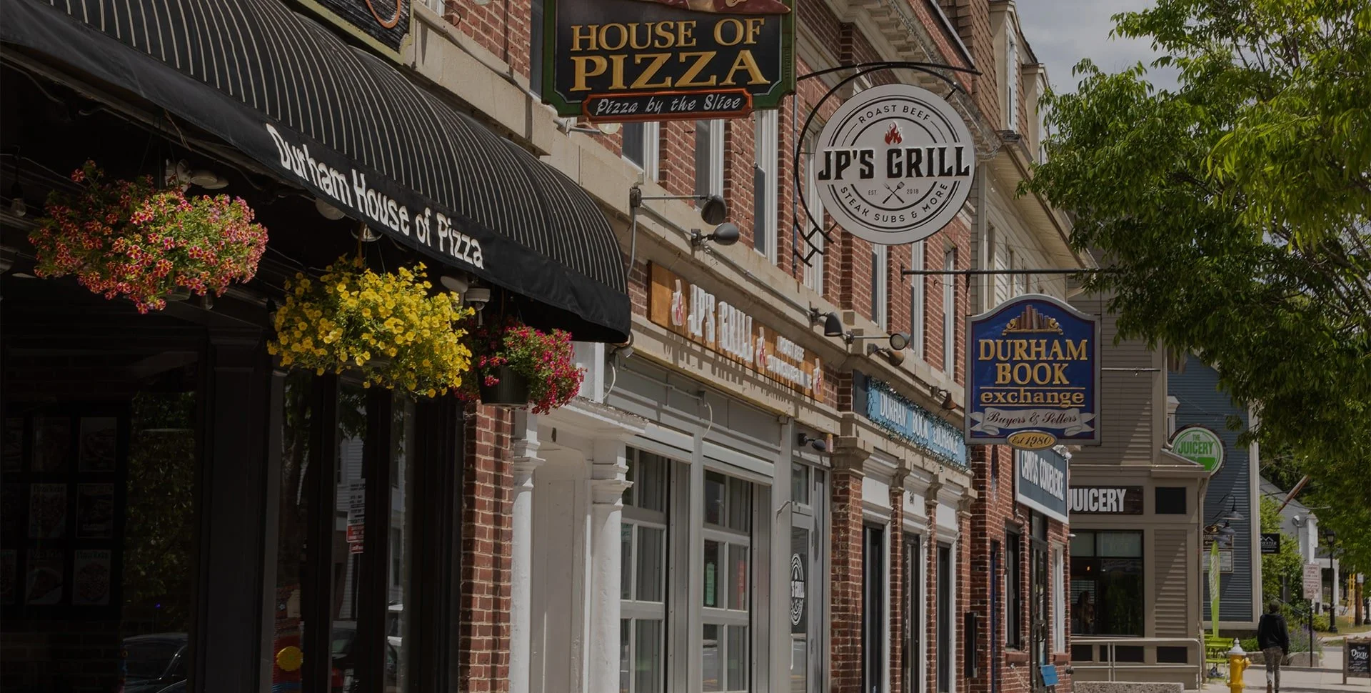 Street view of storefronts with signs for Durham House of Pizza, JP's Grill, Durham Book Exchange, and The Juicery, with hanging flower baskets and trees.