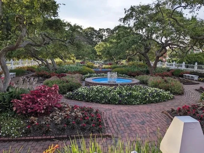 A peaceful garden with a central blue fountain surrounded by colorful flowers and green shrubs, framed by trees with sprawling branches and a white picket fence in the background.