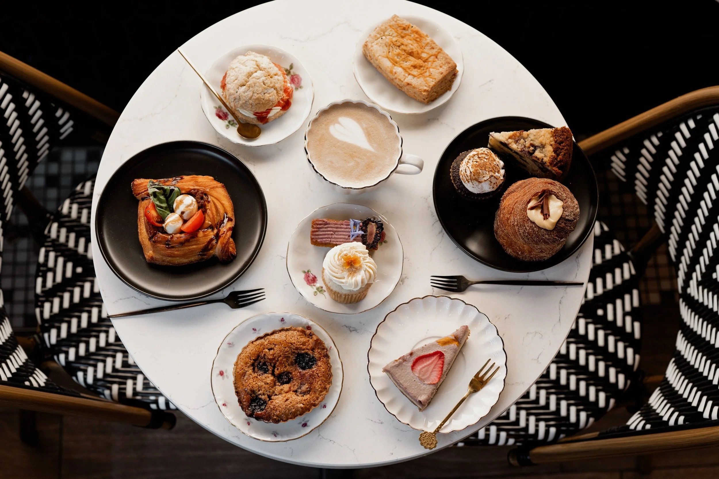 A white round table filled with assorted desserts including cake slices, cookies, a cupcake, and a coffee drink, surrounded by black and white patterned chairs.