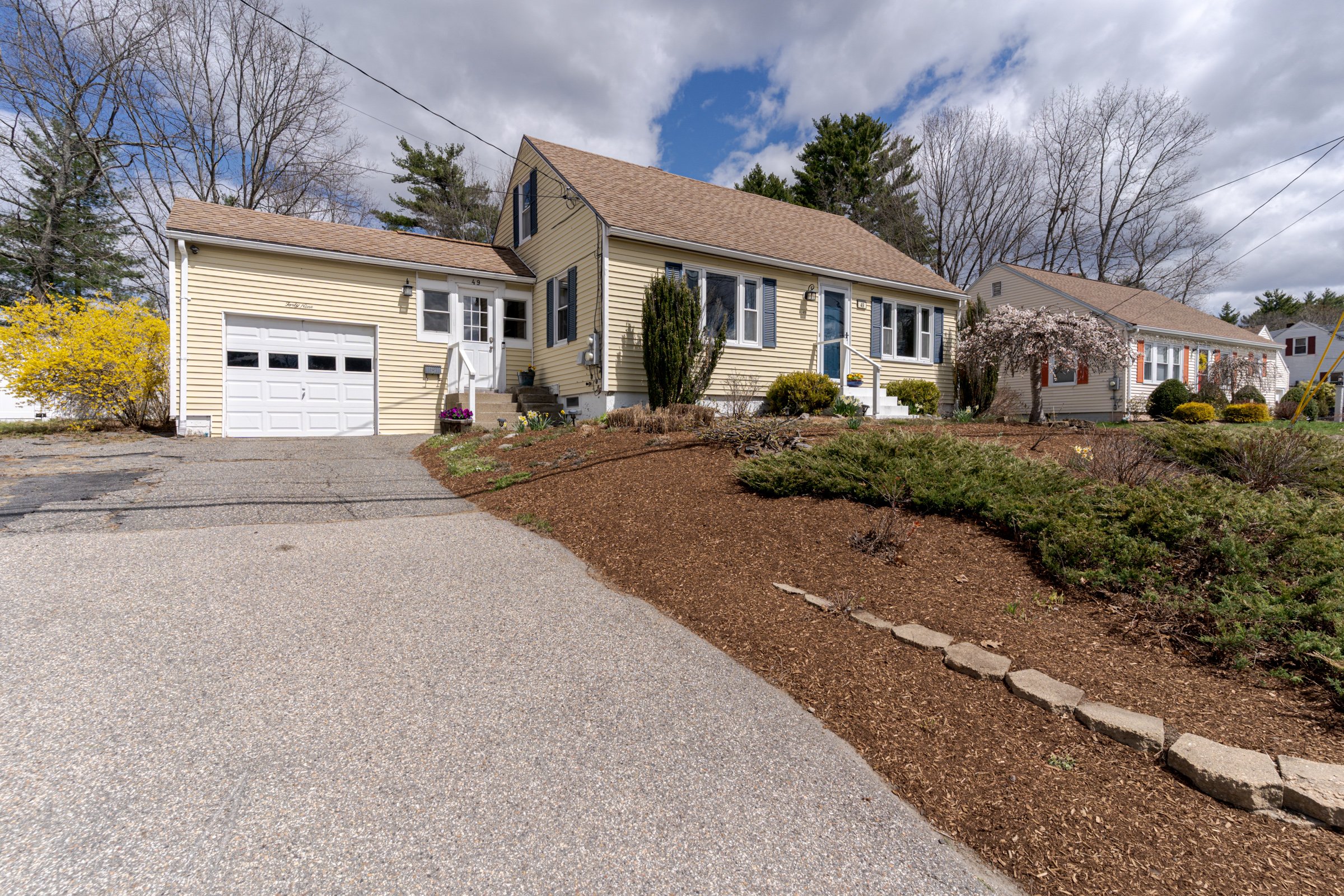 A suburban house with a beige exterior, brown roof, and a one-car garage. The front yard has a small plants and trees, some with blossoms. The driveway is paved, and the sky is partly cloudy.