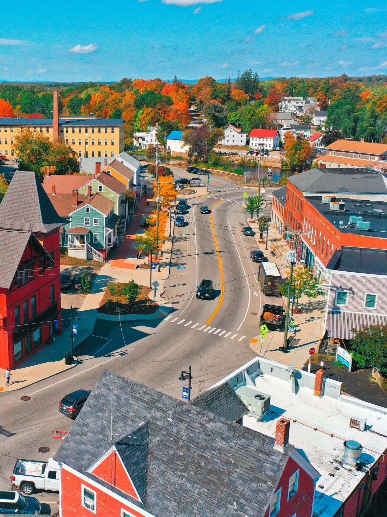 An aerial view of a small town with colorful houses, a main street with cars, a parking lot, and trees with fall foliage under a partly cloudy sky.