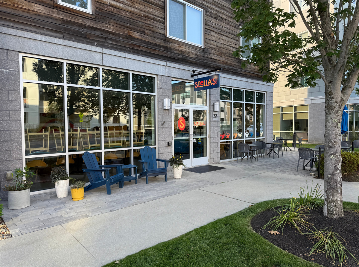 Exterior view of a cafe or restaurant with outdoor seating, large windows, potted plants, and a sign that says 'Stella's'.