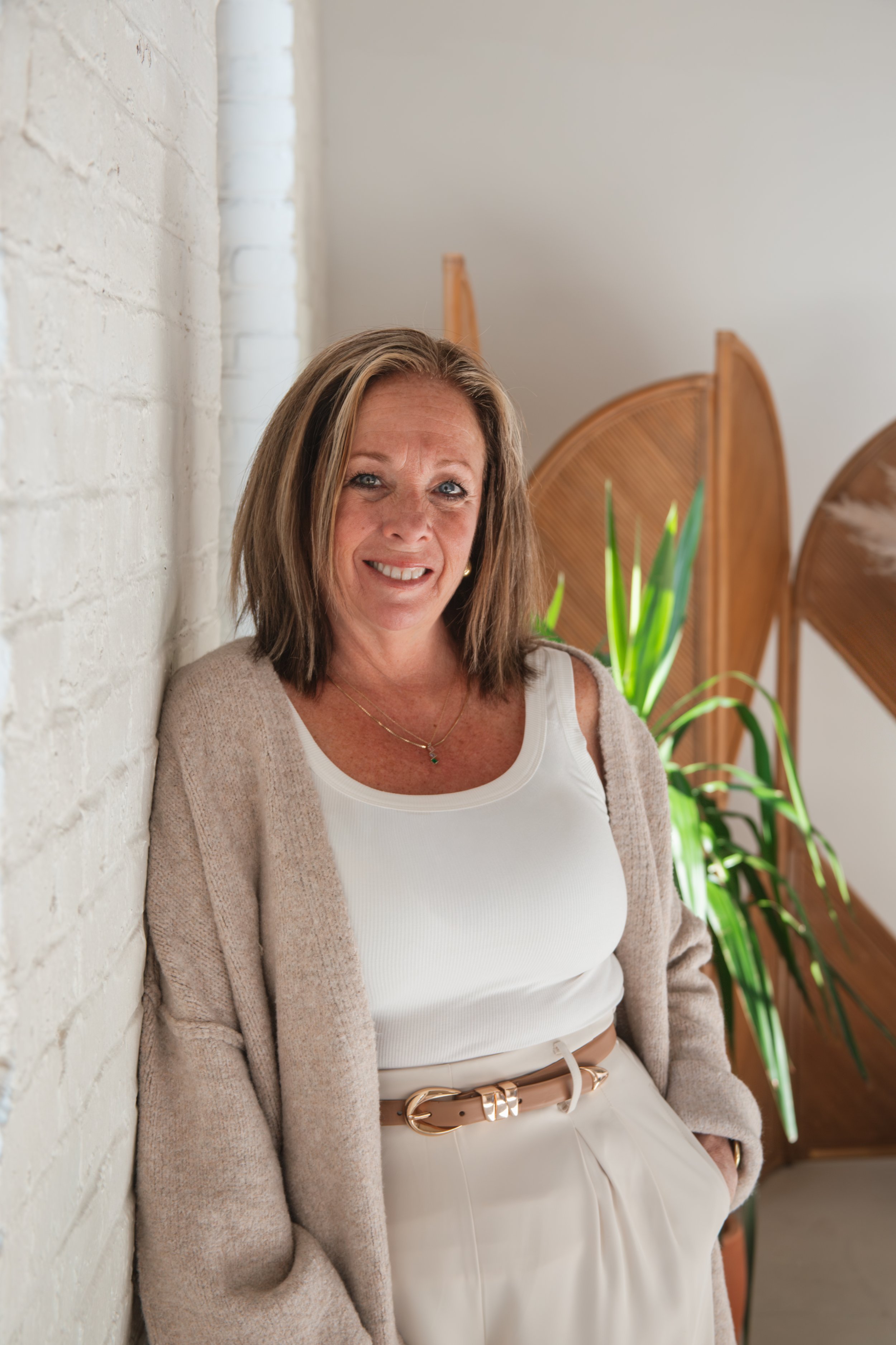 A woman with shoulder-length brown hair, wearing a white tank top, a beige cardigan, and white pants with a gold belt, leaning against a white brick wall with green plants and wooden room dividers in the background.