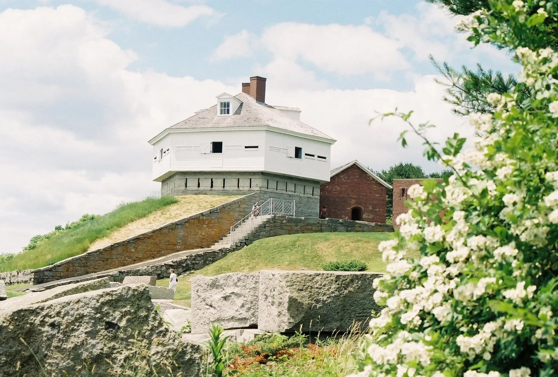 A historic stone lighthouse with a white wooden top, situated on a grassy hill, with a brick building nearby, and people walking up a stone staircase leading to it. There are green trees and blooming bushes in the foreground under a partly cloudy sky.