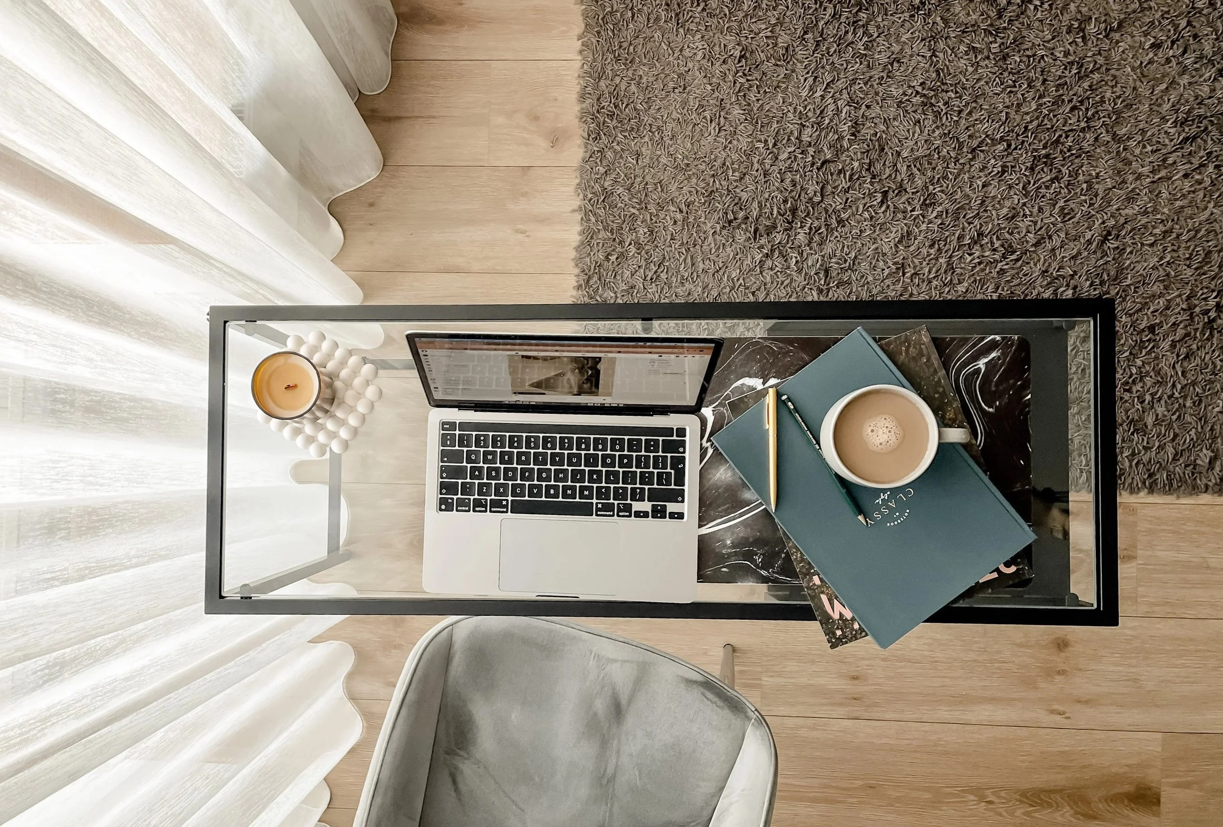 A workspace featuring a glass desk with a laptop, a stack of books, a coffee cup, a candle, and a decorative candle holder, set near a window with sheer curtains.