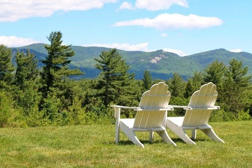 Two white Adirondack chairs facing a scenic view of trees and distant mountains under a partly cloudy sky.