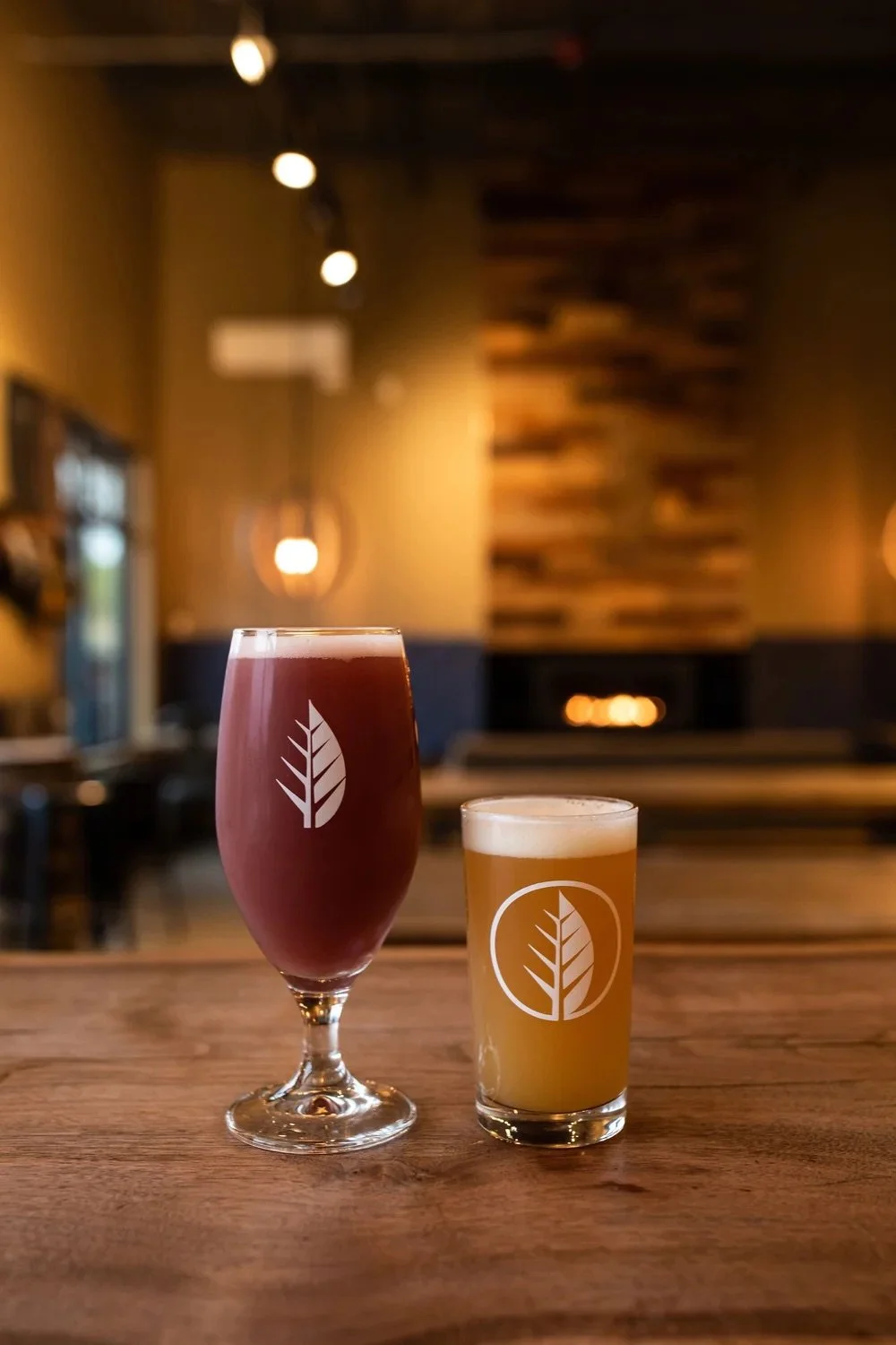 A pink frothy beverage in a tall glass and a pale beer in a short glass with a leaf logo, on a wooden table inside a dimly lit bar or brewery.