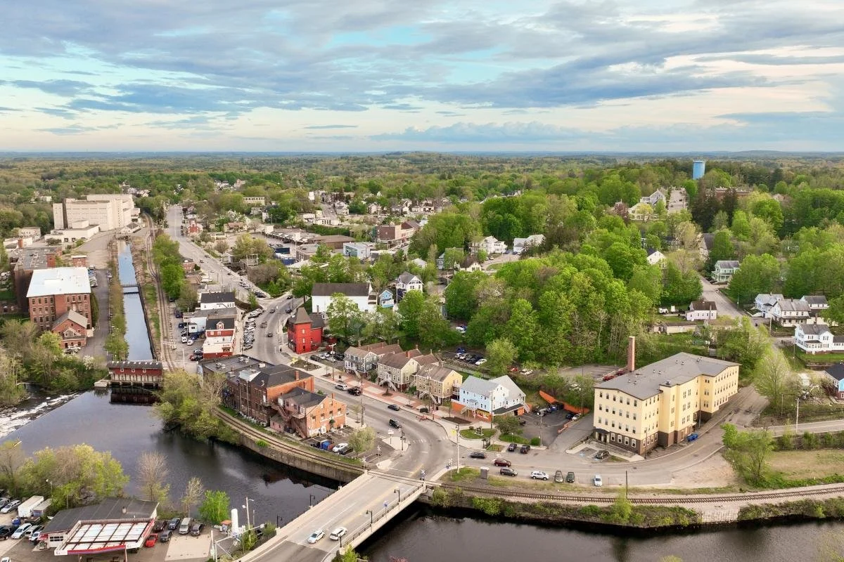 An aerial view of a small town with a river and bridge, surrounded by trees and buildings.