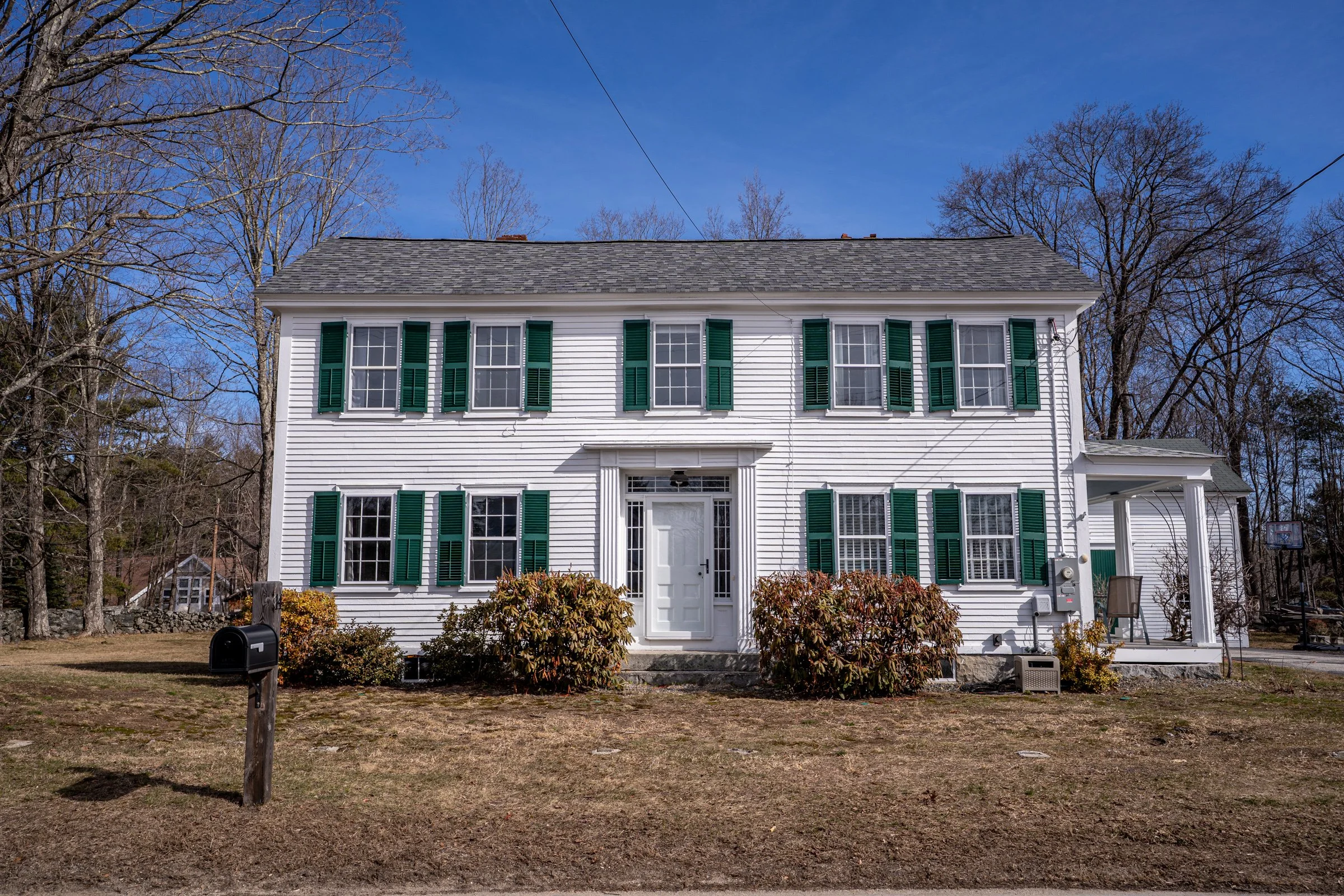 Two-story white house with green shutters under a clear blue sky, surrounded by leafless trees and a front yard with bushes and a mailbox.