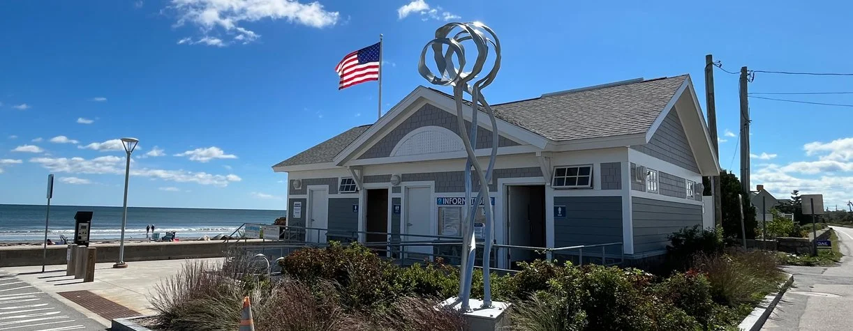 Beachside visitor center with a modern metallic sculpture, American flag, and restrooms, near the ocean under a blue sky.