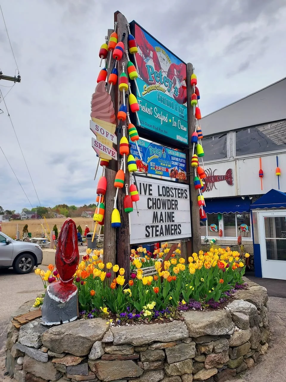 A restaurant sign surrounded by colorful buoys and flowers. The sign advertises seafood such as live lobsters, chowder, Maine, and steamers at Pete's Seafood. There are parking lot and cloudy sky in the background.