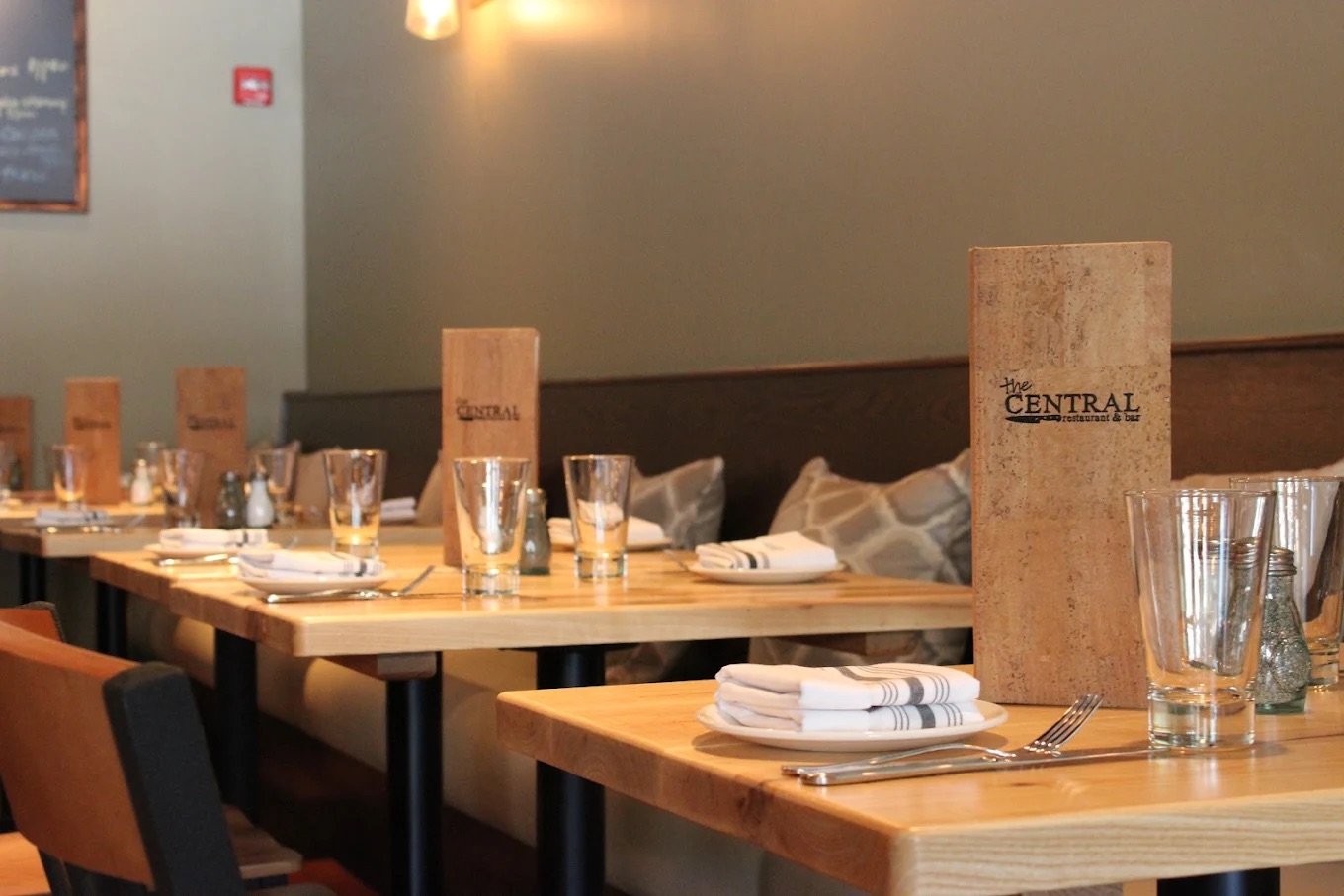Empty restaurant table set with glasses, plates, silverware, cloth napkins, and menu, with a cushioned bench and pillows against the wall.
