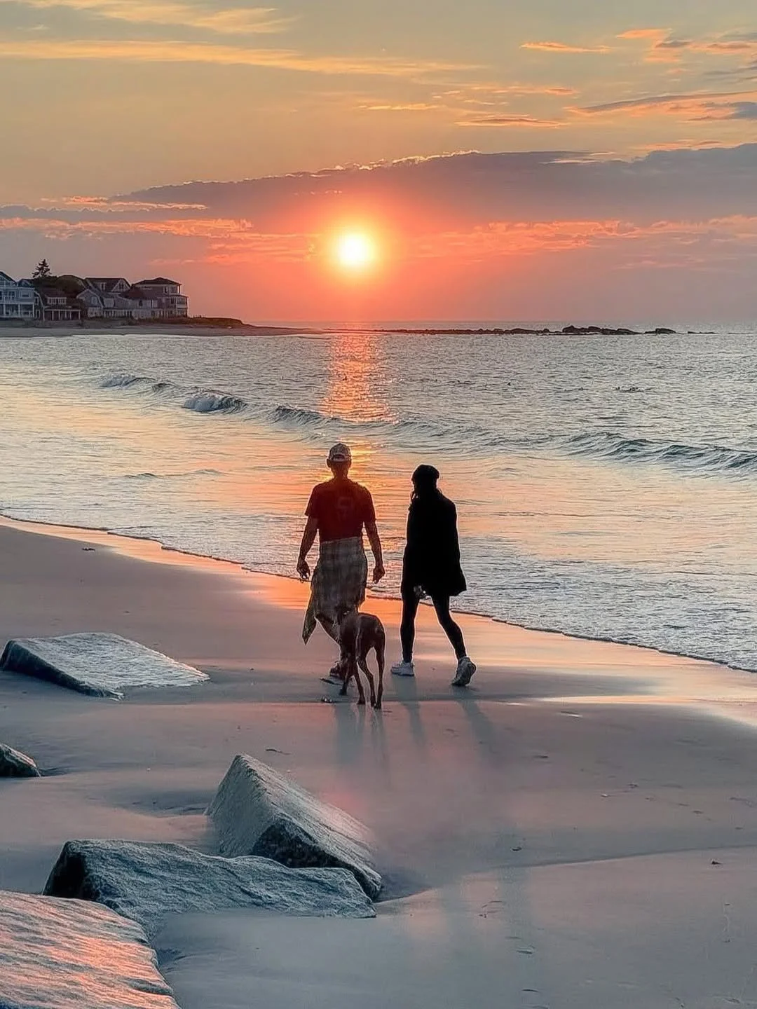 Two people walking with a dog along the beach during sunset, with waves and rocks in the foreground and houses on the distant shoreline.