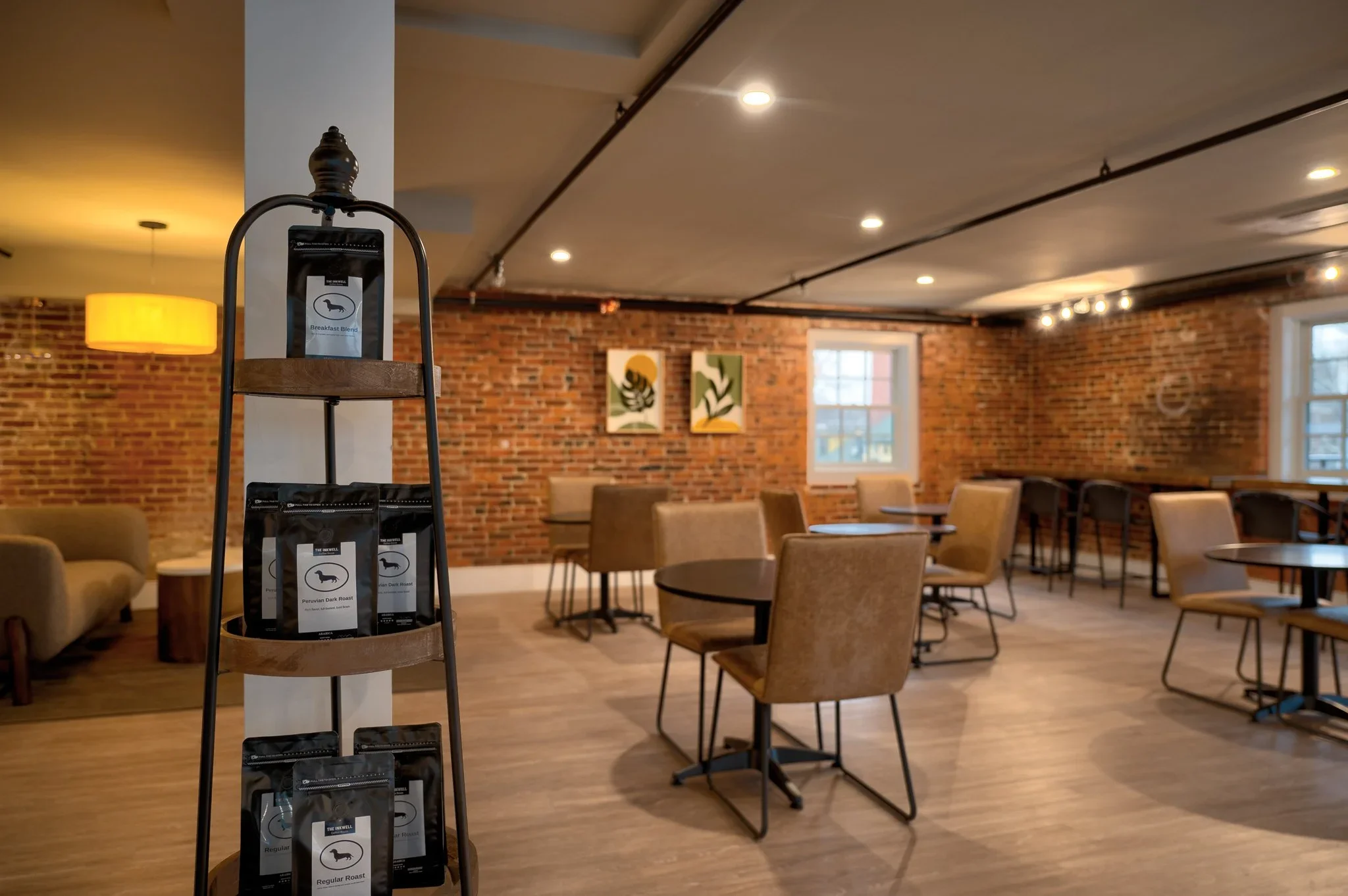 A coffee shop interior with brick walls, large windows, hanging art, and tables with chairs. In the foreground, a display rack holds packets of coffee labeled 'Peruvian Dark Roast.'