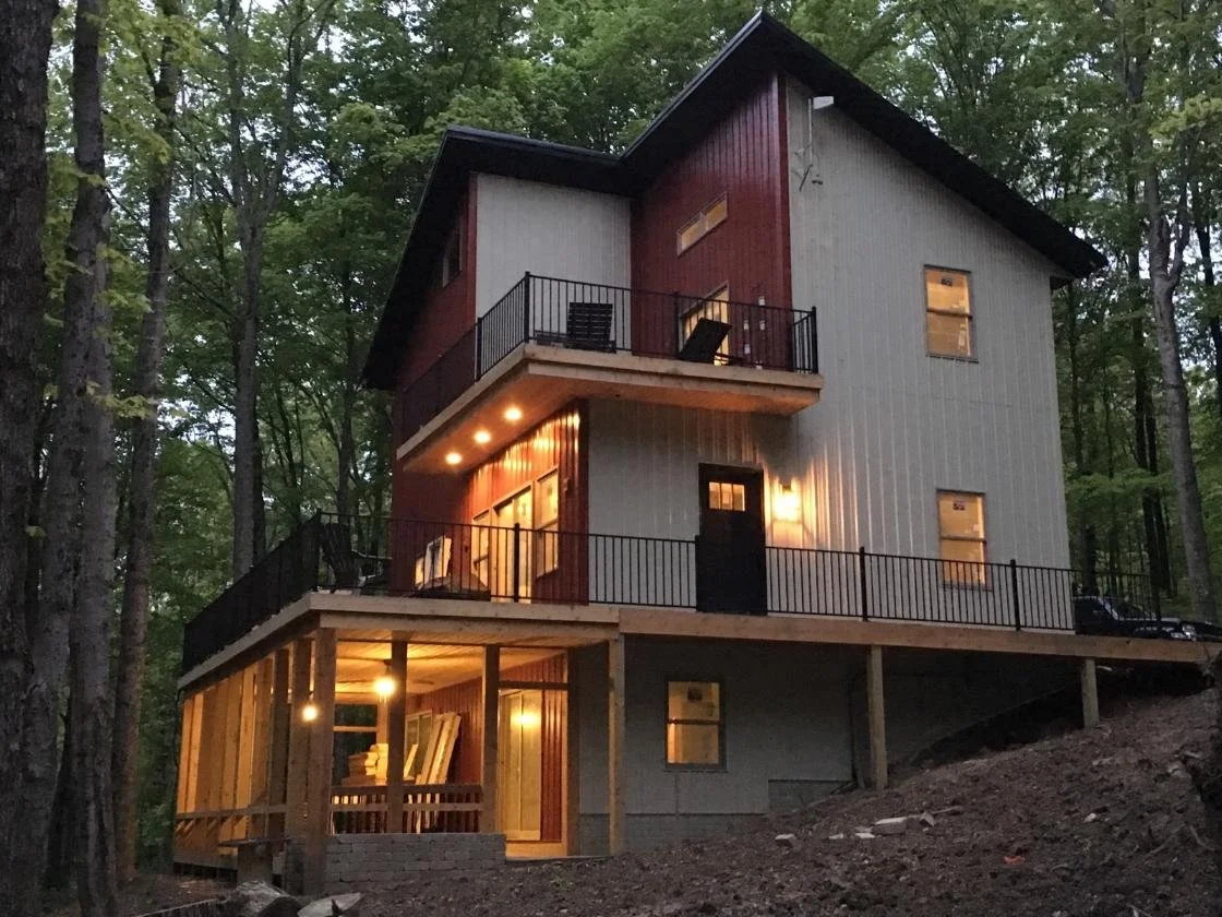 A multi-level house with a combination of red and gray exterior walls, illuminated from inside, surrounded by tall trees, with a wooden deck featuring stunning outdoor lighting. Crafted by Deck Masters - Built with expertise since 1986.