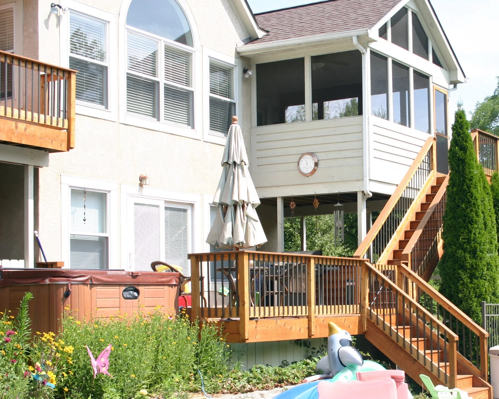 Backyard with wooden deck, patio umbrella, hot tub, children’s inflatable pool, garden, and house with screened porch and multiple windows. Deck Masters has been turning outdoor dreams into reality since 1986.