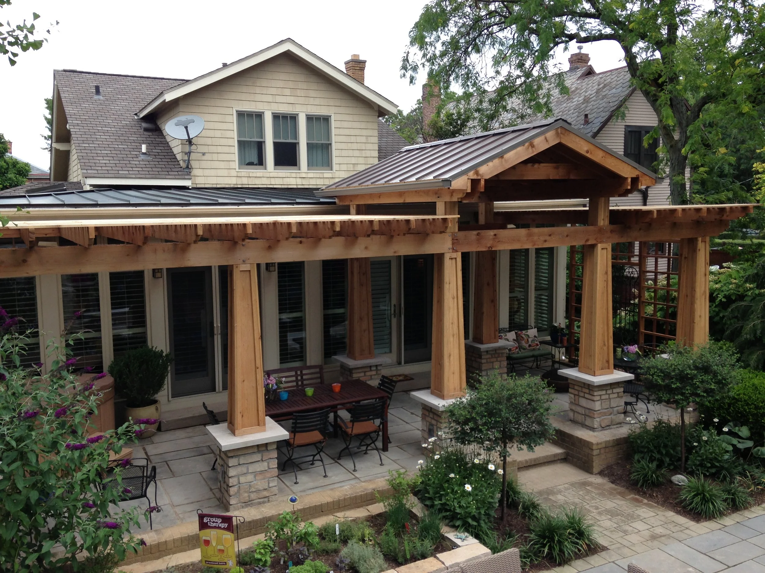 Luxurious wooden pergola, crafted by Deck Masters, with brick pillars over a patio with outdoor furniture, surrounded by a garden with plants and flowers.
