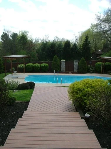 View of a backyard with a wooden deck leading to a swimming pool, surrounded by green bushes and trees. Deck Masters is built on experience, and trusted for generations. Our craftsmanship reflects who we are.