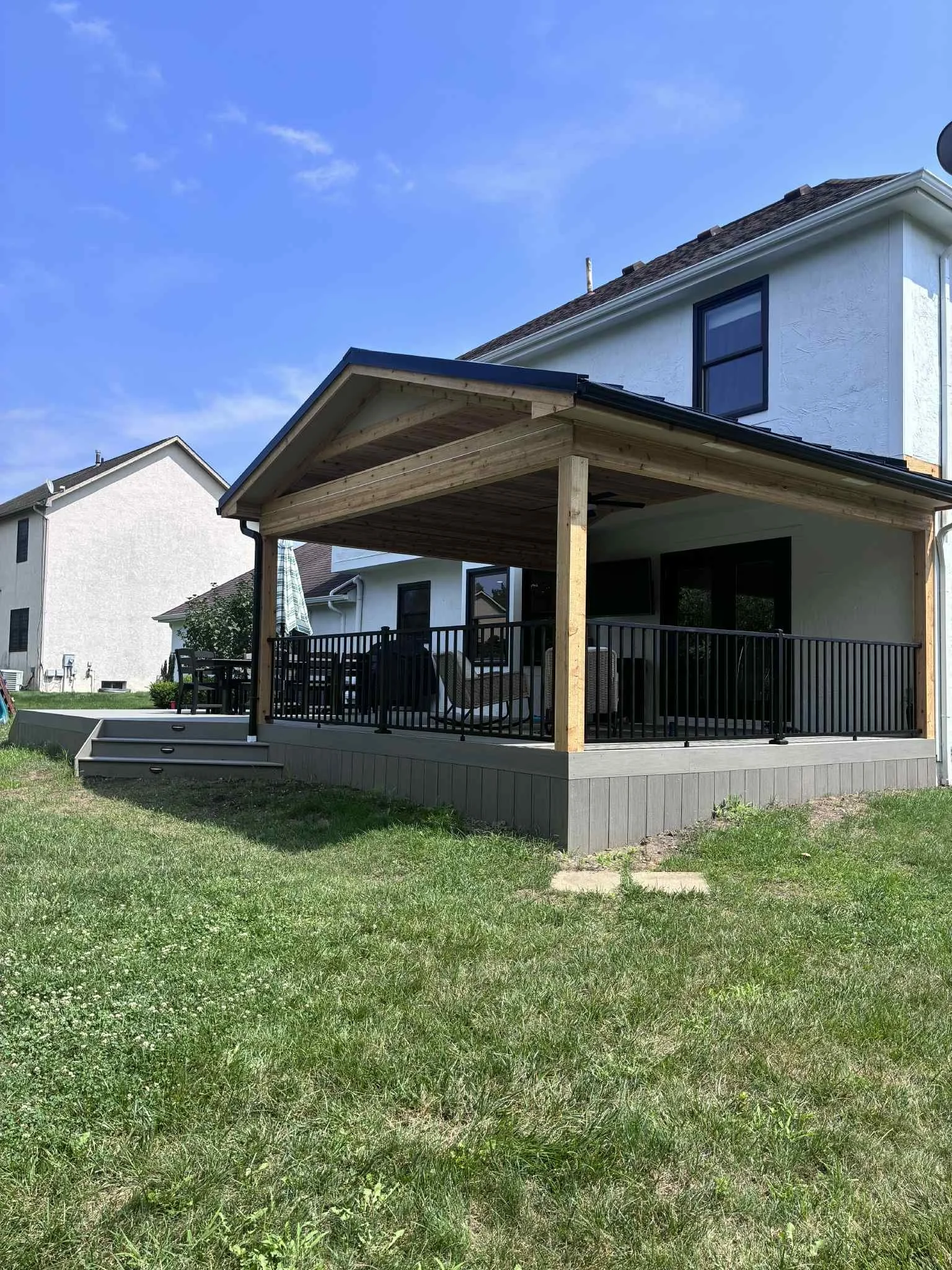 Backyard with a newly constructed wooden and metal railing deck, crafted by expert carpenters at Deck Masters, attached to a white two-story house, with steps leading down to grass and a clear blue sky. Deck Masters is located in Columbus Ohio.