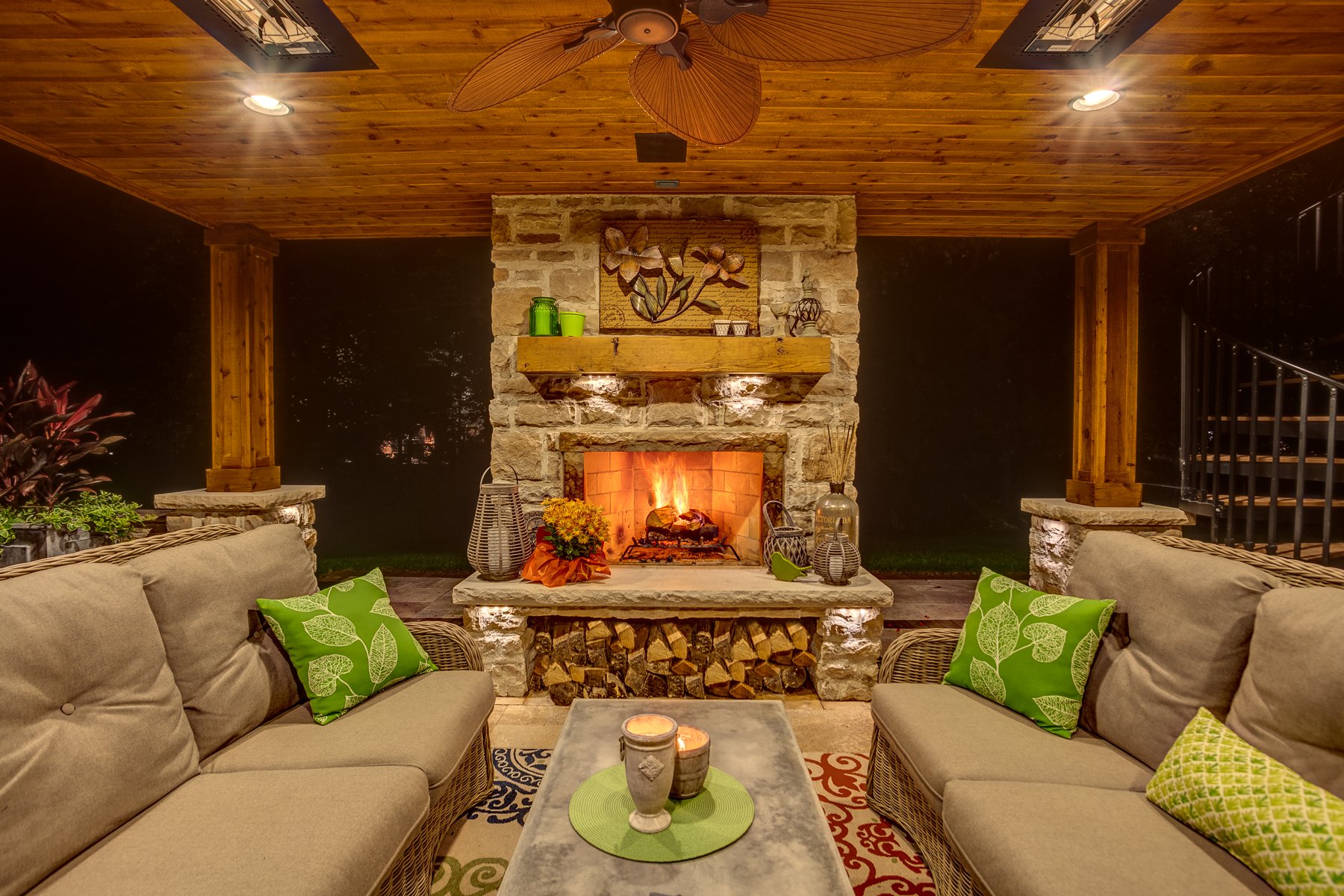 Outdoor living room with a stone fireplace, beige cushioned sofas with green and patterned pillows, a concrete coffee table with candles, and a wooden ceiling with a ceiling fan. Firewood is stacked below the fireplace, and the mantel is decorated with vases and a painting of flowers.