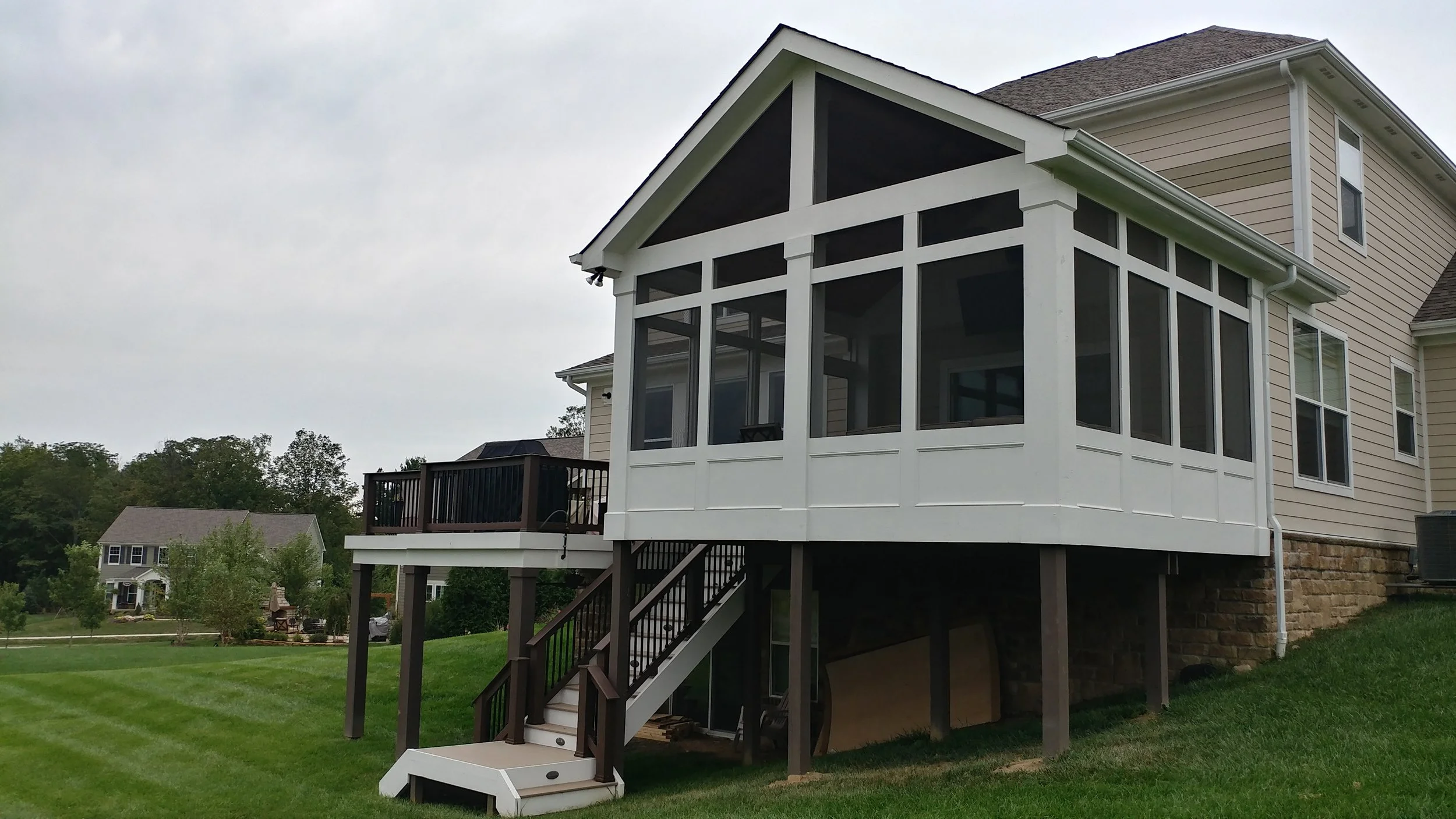 Back view of a two-story house with a screened-in porch and stairs leading to the yard, green grass, neighboring houses in the background, and a cloudy sky.