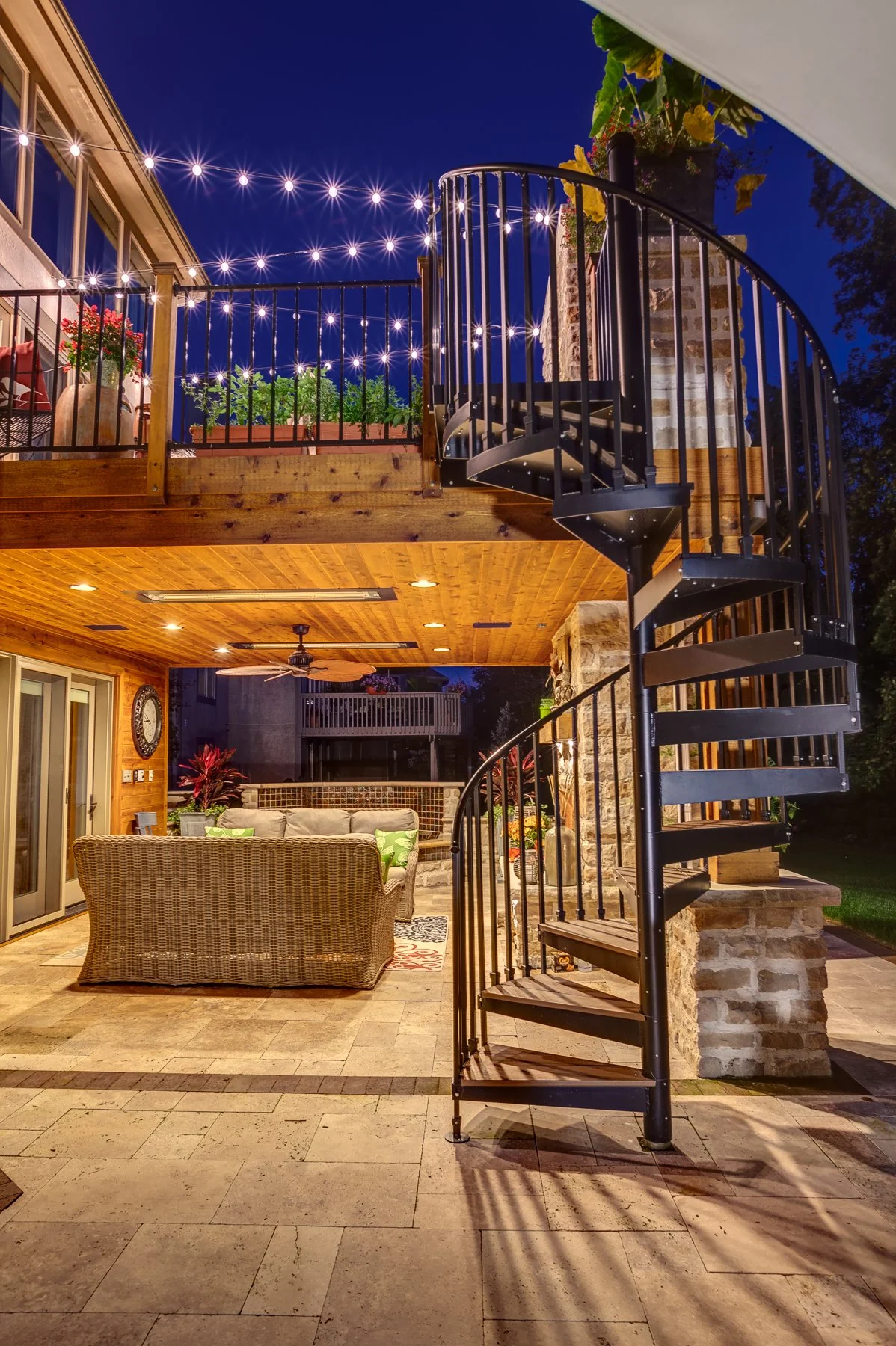 Backyard patio and deck built by Deck Masters in Westerville, Ohio, at night with wicker furniture, potted plants, string lights, and a spiral staircase leading to an upper deck with more plants and outdoor decor.