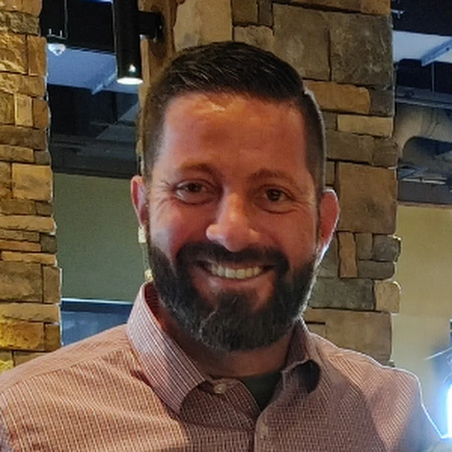 A smiling man with a beard and short hair wearing a checkered shirt inside a restaurant with stone walls and a window.